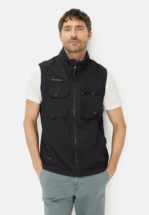 Mann trägt schwarze Utility-Weste mit Reißverschluss über weißem Kurzarmhemd und hellgrauer Hose, steht mit Händen in den Taschen.