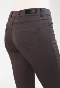 Tmavě hnědé džíny z denimu s úzkým střihem, zadními kapsami a koženou etiketou na pásku. Tkanina se zdá být hladká a elastická.