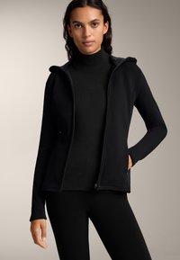 Črna zip-up dukserica z mehkim, flisastim oblogom znotraj. Dolgi rokavi in žepi, zasnovana za oprijet videz. Nosi se čez črn pulover s stoječim ovratnikom.