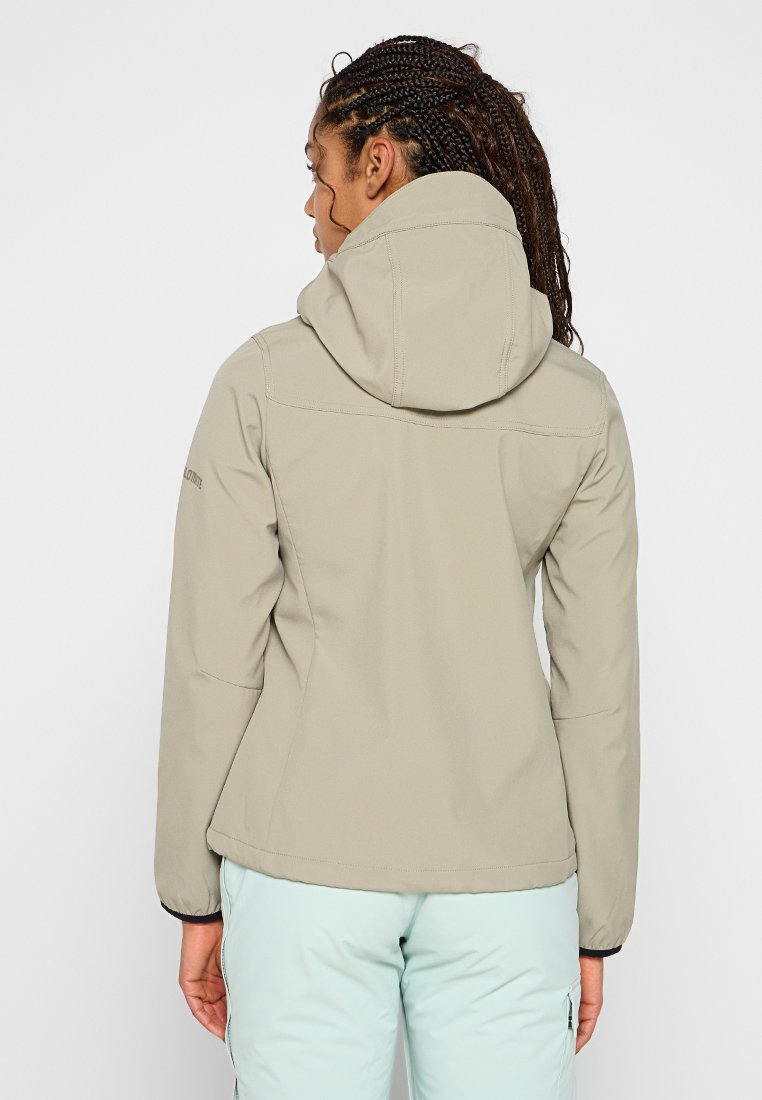Donna con capelli intrecciati che indossa una giacca con cappuccio beige e pantaloni blu chiaro, in piedi di spalle contro uno sfondo neutro.