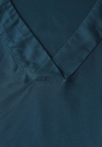 Tessuto satinato turchese con scollatura a V. Texture liscia e leggero lucido. Cuciture ordinate lungo il colletto e le spalle.