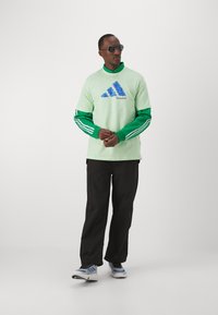 T-shirt verde e menta con logo Adidas blu, sovrapposto a una maglietta a maniche lunghe verde. Pantaloni neri e sneakers multicolori completano l'outfit.