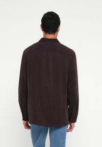 Sötétlila, hosszú ujjú ing gallérral, hátul redővel és gombos mandzsettával, világoskék farmernadrággal párosítva. A textúra puha érzetű.