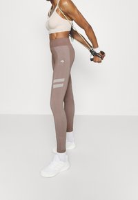 Brązowe legginsy sportowe z wysokim stanem, z odblaskowymi paskami i kontrastowymi szwami, zestawione z jasnym stanikiem sportowym i białymi sneakersami.