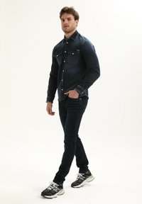 Donkerdenim shirt met twee borstzakken, knoopsluiting en contrasterende stiksels, gecombineerd met een donkere spijkerbroek en gestructureerde sportschoenen.