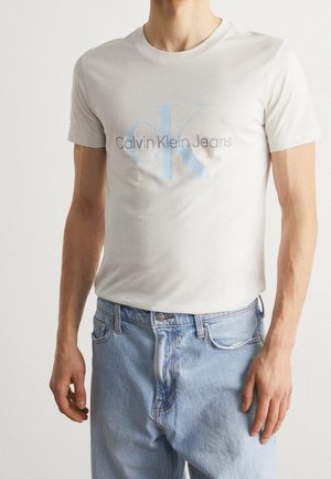 Mand iført en lysegrå Calvin Klein Jeans t-shirt og lyseblå jeans, stående mod en ensfarvet baggrund.