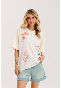 Krémové tričko s krátkym rukávom a farebnými potlačami na západný motív, vrátane koní, kaktusov a lebky, skombinované so svetlými džínsovými šortkami.