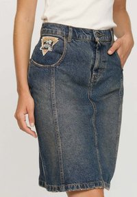 Jupe en denim bleu foncé, avec une poche frontale ornée d'un patch logo, d'une coupe mi-longue et d'un ourlet brut. Détails de couture visibles sur l'ensemble.