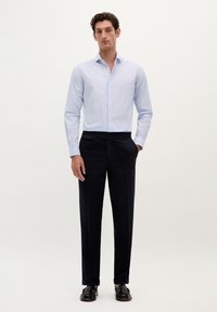 Camisa de botones azul claro con botones blancos, combinada con pantalones oscuros de color navy y zapatos negros. Tela suave, corte ajustado, diseño minimalista.