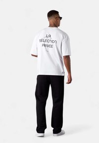 White short-sleeve t-shirt featuring bold black text on the back. Made of cotton, relaxed fit, worn with black trousers and white sneakers.