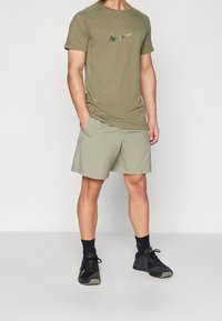 Camisa de manga corta verde oliva con un logotipo de Nike texturizado, combinada con shorts verdes claros y zapatillas deportivas negras, de pie sobre un fondo blanco.