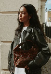 Kobieta z długimi ciemnymi włosami, ubrana w czarną skórzaną kurtkę i białą koszulę, trzyma brązową torbę w czarne plamki, stojąc na świeżym powietrzu przy ścianie.