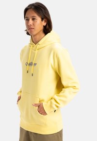 Sudadera amarilla con capucha, con un bolsillo frontal y cordones. Presenta un logotipo gráfico en azul marino en el pecho. Tejido suave y texturizado.