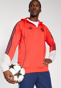 Sweat à capuche de sport rouge avec des rayures bleu marine, col zippé, accents de poignets blancs et un logo Adidas, accompagné d'un ballon de football noir et blanc.