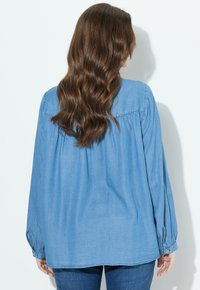 Donna con lunghi capelli castani ondulati, che indossa una camicia di jeans azzurro chiaro a maniche lunghe e jeans blu scuro, in piedi contro uno sfondo bianco.