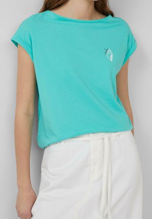 Femme portant un haut turquoise à manches courtes avec un petit motif de coquillage blanc et un pantalon blanc taille haute avec une ceinture nouée.