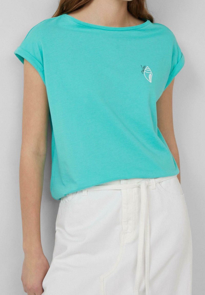 Kvinna som bär en turkos kortärmad topp med en liten vit snäcka-design och höga vita byxor med ett knytbandsbälte.