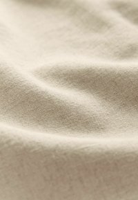 Lichtbeige linnen stof met een gestructureerd oppervlak, dat zachte plooien en subtiele variaties in kleur en textuur over het materiaal vertoont.