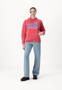 Rød hettegenser med blå grafisk tekst, frontlomme og snor. Kombinert med lyseblå vide jeans og svarte sko. Speilsolbriller brukes.