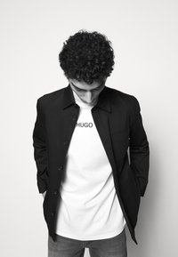 Jeune homme aux cheveux bouclés regardant vers le bas, portant une veste noire boutonnée par-dessus un T-shirt blanc avec le texte « HUGO », et un jean bleu.