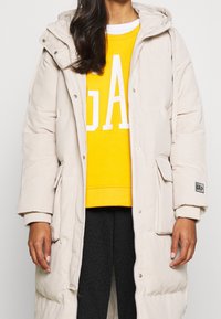 Persona che indossa un lungo piumino beige sopra una felpa gialla GAP con pantaloni neri, in piedi davanti a uno sfondo bianco uniforme.