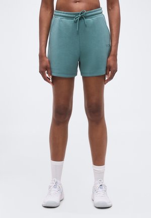 Personne portant un short turquoise à cordon, des chaussettes montantes blanches et des chaussures de sport blanches, debout devant un fond clair uni.