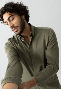 Maglietta verde oliva a maniche lunghe con bottoni, caratterizzata da una texture liscia e maniche arrotolate, indossata da una persona mentre regola il proprio braccio.