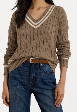 Femme portant un pull à col en V marron à torsades avec des rayures blanches, rentré dans un jean bleu avec une ceinture marron.
