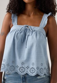 Light blue sleeveless top in soft cotton with ruffled straps, a tie front, and eyelet trim featuring floral cutouts along the hem.