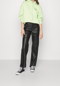 Persoon met een limegroene hoodie, zwarte rechte broek van nep leer en zwarte hoge sneakers, staand tegen een witte achtergrond.