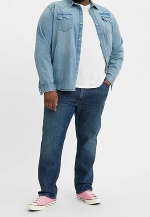 Mężczyzna w niebieskiej denimowej koszuli na białym t-shircie, niebieskich dżinsach i różowych płóciennych trampkach stoi z jedną ręką w kieszeni.