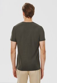 Ciemnozielona koszulka z krótkim rękawem, w stylu z okrągłym dekoltem, o miękkiej teksturze, prostej nowoczesnej kroju, widziana z tyłu, w połączeniu z jasnymi spodniami.