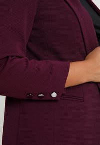 Bordeaux blazer met ribbelstructuur en zilveren knopenaccenten op de mouwen, met een aansluitende pasvorm en gestructureerde revers.