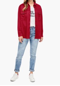Kvinde iført en rød skjorte med knaplukning over en hvid grafisk t-shirt, lyseblå mønstrede jeans rullet op ved anklerne, og hvide sneakers.
