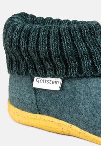 Ciemnozielony filcowy pantofel z prążkowanym dzianinowym mankietem, żółtą podeszwą i małą etykietą "Gottstein" z boku. Teksturowane wykończenie materiału.