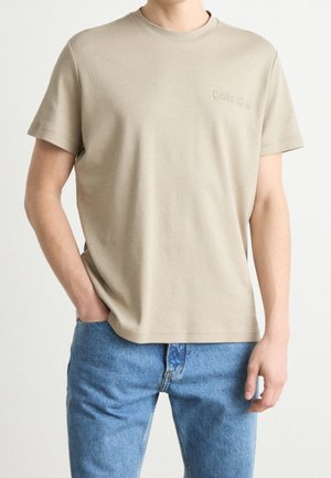 T-shirt beige à manches courtes avec un logo subtil "Calvin Klein" sur la poitrine, porté avec un jean bleu clair, main dans la poche, fond neutre.