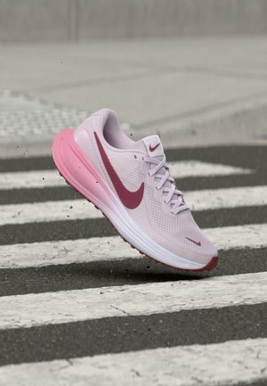 Ανοιχτό ροζ αθλητικό παπούτσι Nike με σκούρο ροζ σήμα και σόλα, αιωρείται πάνω από διαβάση πεζών σε ασφαλτοστρωμένο δρόμο.