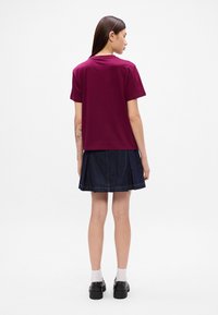 Burgunderfarget bomull T-skjorte med korte ermer, kombinert med en mørk denim plissert skjørt. Modellen har på seg svarte sko og hvite sokker.