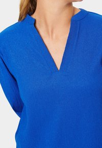 Blusa blu strutturata con scollatura a V, caratterizzata da sottili righe verticali. Tessuto morbido con un leggero lucido.