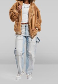 Brązowa futrzana kurtka na zamek z przodu, biała dopasowana koszula, jasnoniebieskie wysokowciskane dżinsy oraz masywne sneakersy z teksturowaną podeszwą.