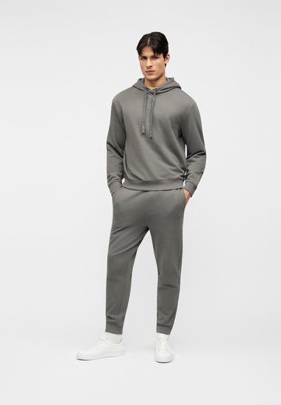 Jeune homme debout, les mains dans les poches, portant un sweat à capuche gris, un pantalon de survêtement assorti, des chaussettes blanches et des baskets blanches, sur un fond uni.