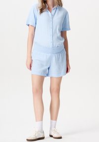 Světle modrý krátký rukáv s knoflíky z texturované látky ve spojení se sladěnými šortkami s elastickým pasem. Bílé tenisky s detaily.