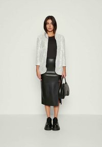 Blazer prateado com lantejoulas sobre uma blusa preta, combinado com uma saia midi de couro preta com fendas laterais e ténis pretos; a segurar uma mala preta.