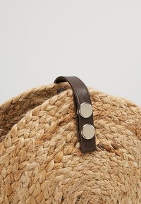 Rund flettet stråtaske med mørkebrunt læderstrop med to sølvknapper på en lys baggrund.