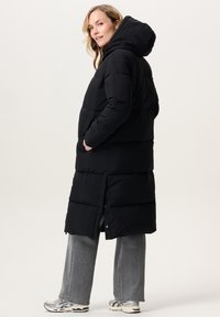 Vrouw in een lange zwarte pufferjas met capuchon, grijze wijde spijkerbroek en witte sneakers, staand in zij-aanzicht met handen in de zakken.