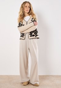Vrouw met lang blond haar, gekleed in beige wijde broek, een crèmekleurige cardigan met zwarte borduursels en tan schoenen, staande tegen een effen muur.