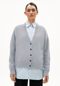 Hellgrauer Cardigan mit V-Ausschnitt, übergroßen Ärmeln, sechs schwarzen Knöpfen und gerippten Bündchen, über einem hellblauen Hemd getragen.