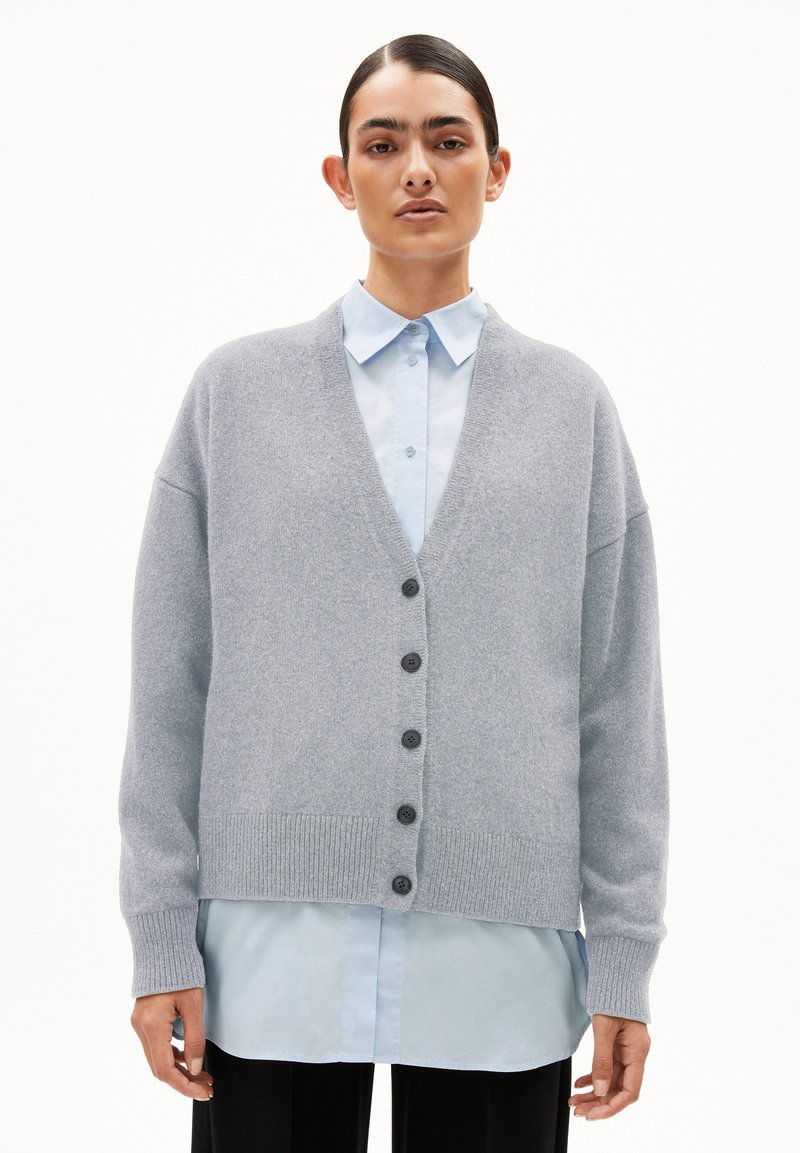 Hellgrauer Cardigan mit V-Ausschnitt, übergroßen Ärmeln, sechs schwarzen Knöpfen und gerippten Bündchen, über einem hellblauen Hemd getragen.