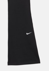 Melnās Nike sporta legingi ar mazu baltu Nike swoosh logotipu tuvumā ceļgala uz baltas fona.