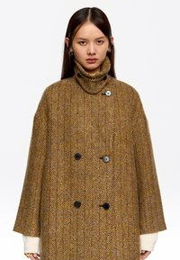 Abrigo marrón de espiga con doble botón, cuello alto y mangas anchas; hecho de tejido texturizado, con sutiles acentos en negro.